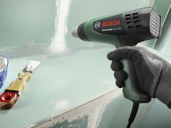 Mano guantata impugna pistola termica Bosch per lavori di finitura. Spatola e stucco a lato.