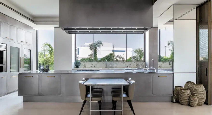 Cucina moderna con isola centrale in acciaio opaco, piano in marmo, tavolo e sedie. Ampia finestra con vista sul verde.