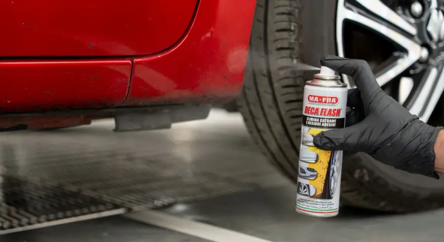 Mano con guanto nero spruzza spray "Deca Flash" su un'auto rossa per come sciogliere la colla secca e residui adesivi.