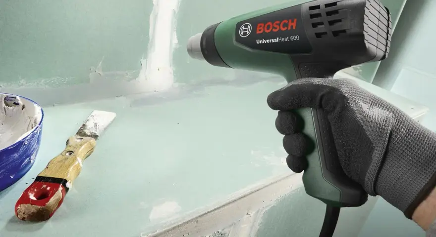 Mano guantata impugna pistola termica Bosch per lavori di finitura. Spatola e stucco a lato.
