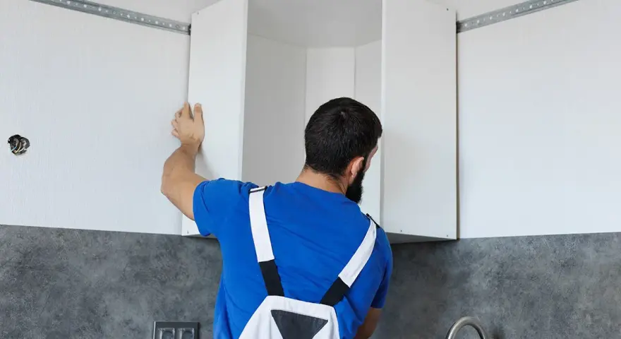 Installazione di pensili da cucina: un uomo con tuta blu monta un pensile angolare su una guida a muro.