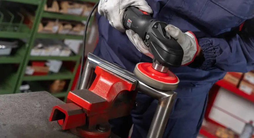 Operaio usa una lucidatrice con dischi abrasivi, parte dei prodotti per lucidare l'acciaio, per rifinire un pezzo metallico fissato in una morsa.