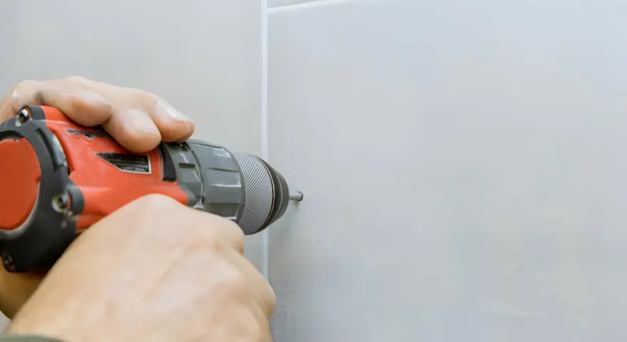 Mani che usano un trapano per fare un buco nel muro bianco.