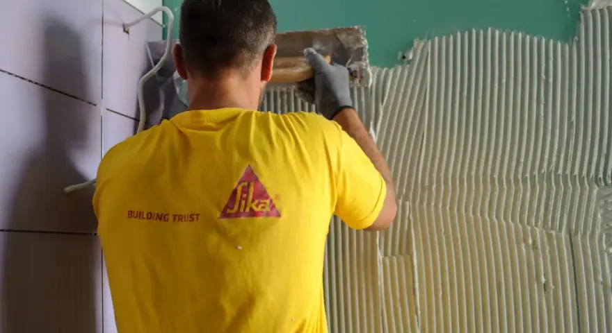 Un operaio con maglia gialla applica colla sul muro, mostrando come attaccare piastrelle.