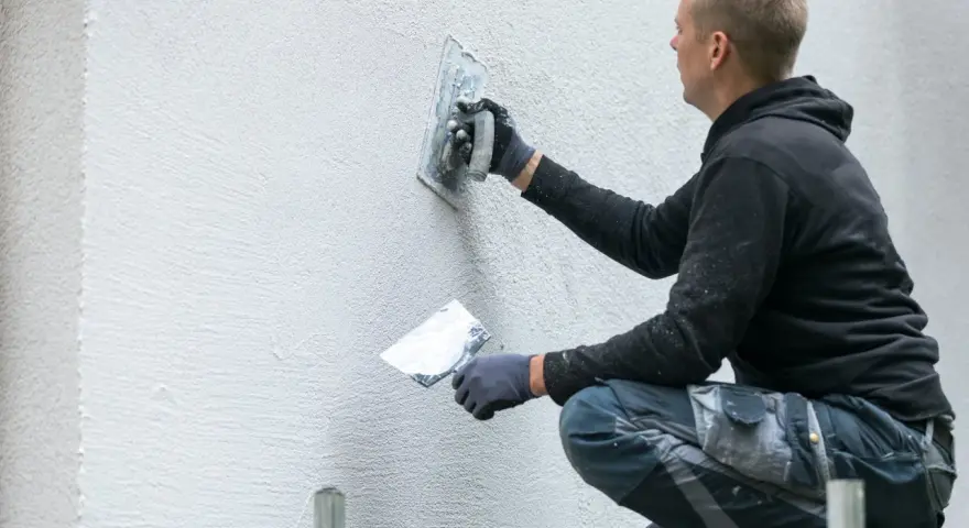 Operaio con cazzuola lavora su un muro esterno, mostrando come rasare un muro rovinato per un aspetto liscio e uniforme.