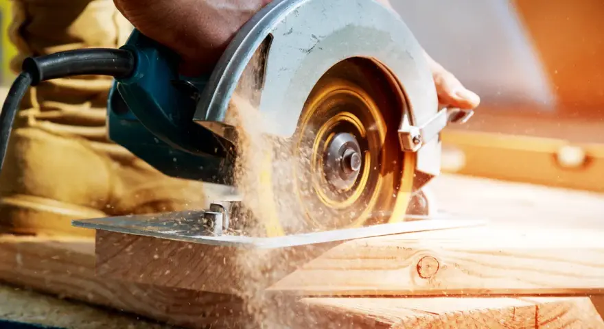 Mano che usa una sega circolare, uno dei tanti tipi di seghe, per tagliare legno. La polvere di legno vola nell'aria.