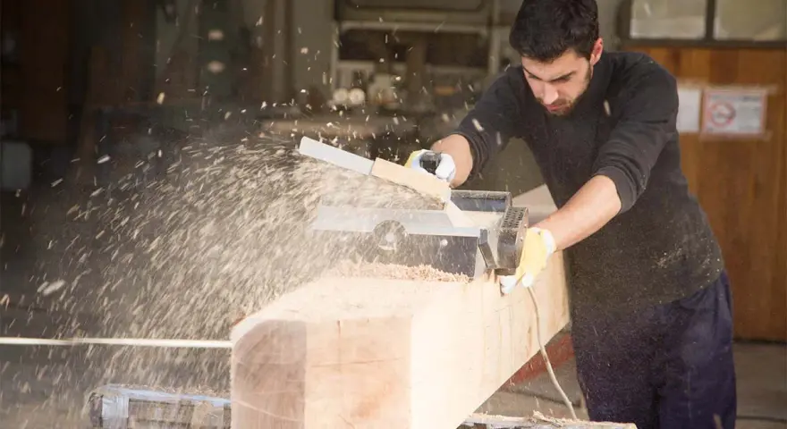 Falegname usa pialla elettrica su un grosso tronco di legno, creando una nuvola di segatura. Il piallare significato è dare una finitura liscia.