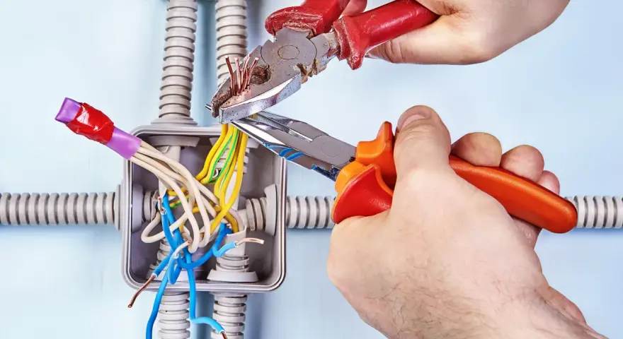 Mani esperte usano **le tenaglie** per lavorare sui fili elettrici in una scatola di derivazione, con tubi corrugati sullo sfondo.