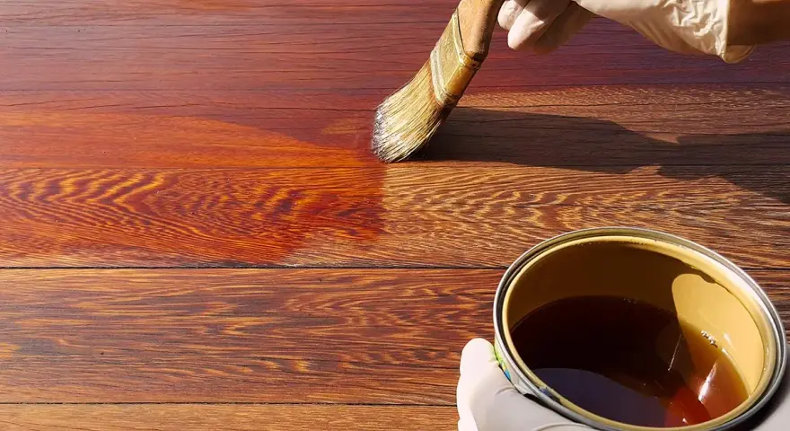 Mano guantata applica una vernice scura su assi di legno, mostrando come scurire il legno per un aspetto più profondo e ricco.