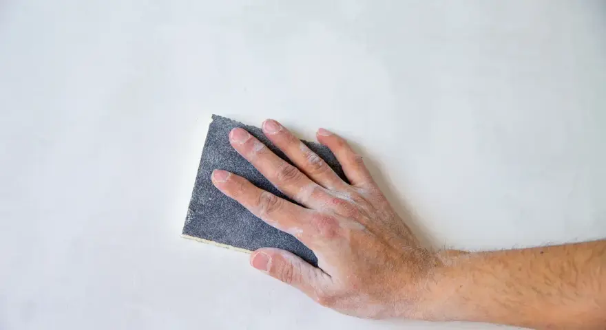 Mano che leviga una parete con carta vetrata, mostrando le sue caratteristiche abrasive per una finitura liscia.