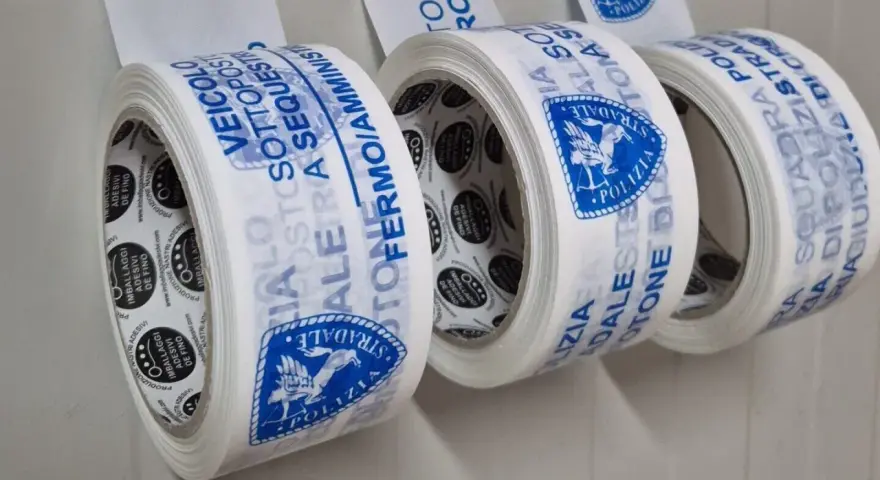 Rotoli di nastri adesivi con scritte blu: "Veicolo Sottosequestro", "Polizia Stradale". Diversi tipi di nastri per imballaggi.