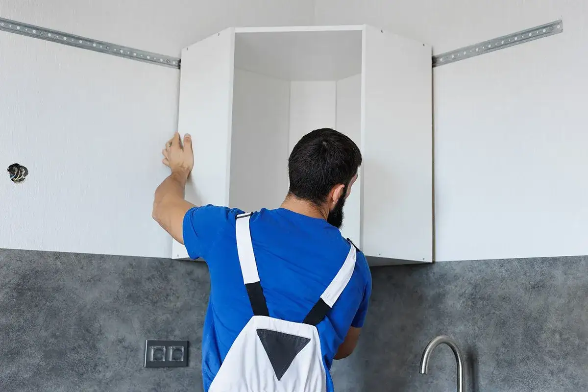 Operaio con tuta blu e bretelle bianche che mostra come appendere i pensili della cucina, fissandoli a una barra metallica.