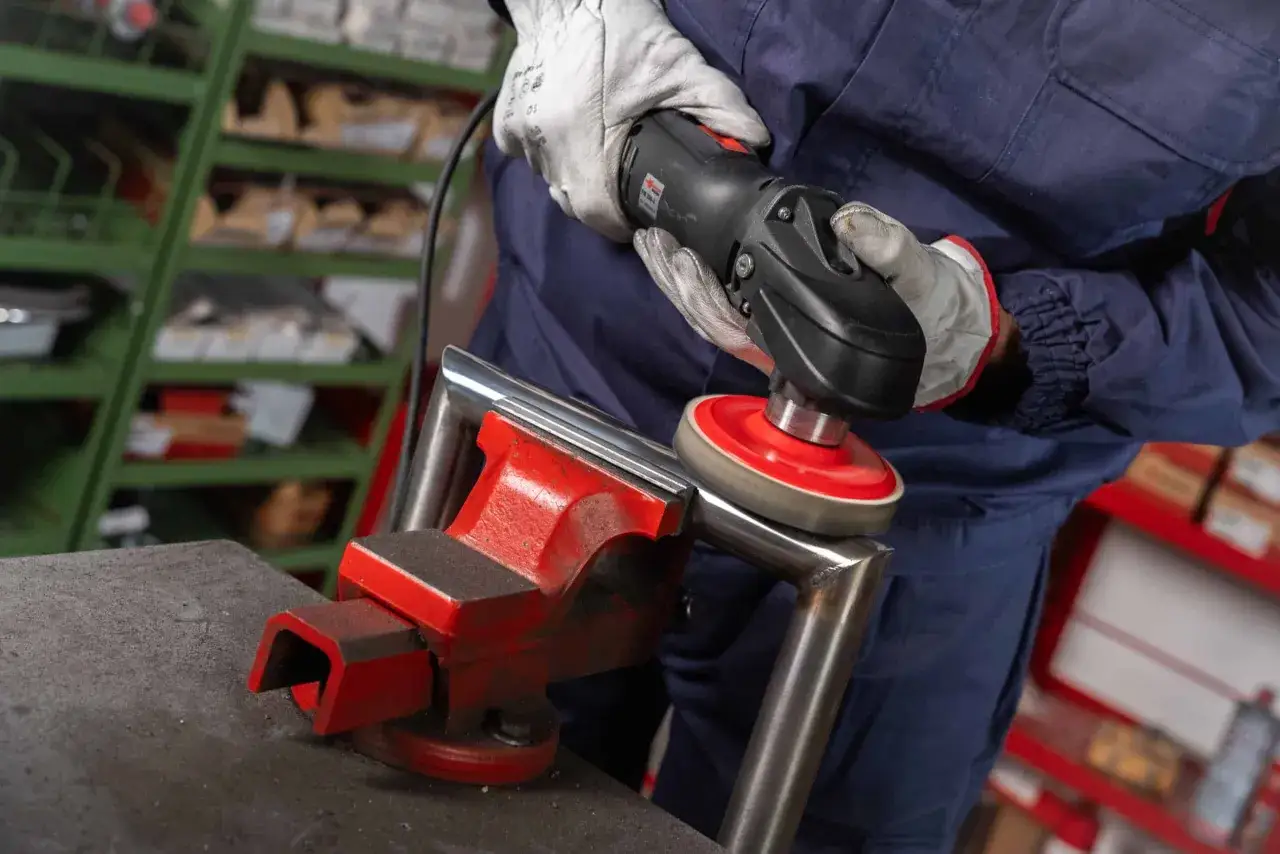 Operaio con guanti lucida un tubo d'acciaio fissato in una morsa, usando un utensile elettrico con disco abrasivo. Prodotti per lucidare l'acciaio in uso.
