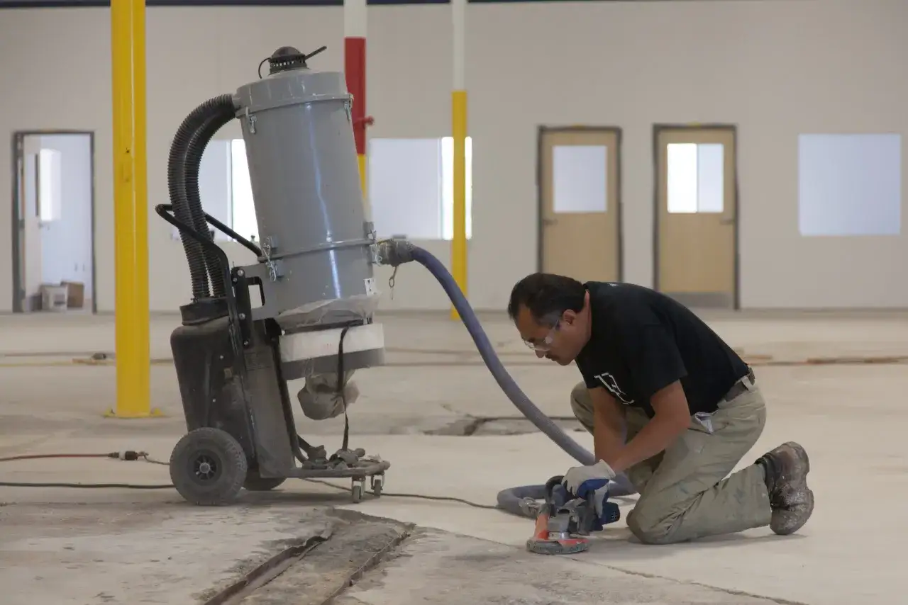 Operaio lavora su un pavimento in resina, utilizzando una smerigliatrice collegata a un aspiratore industriale per rimuovere detriti e polvere.