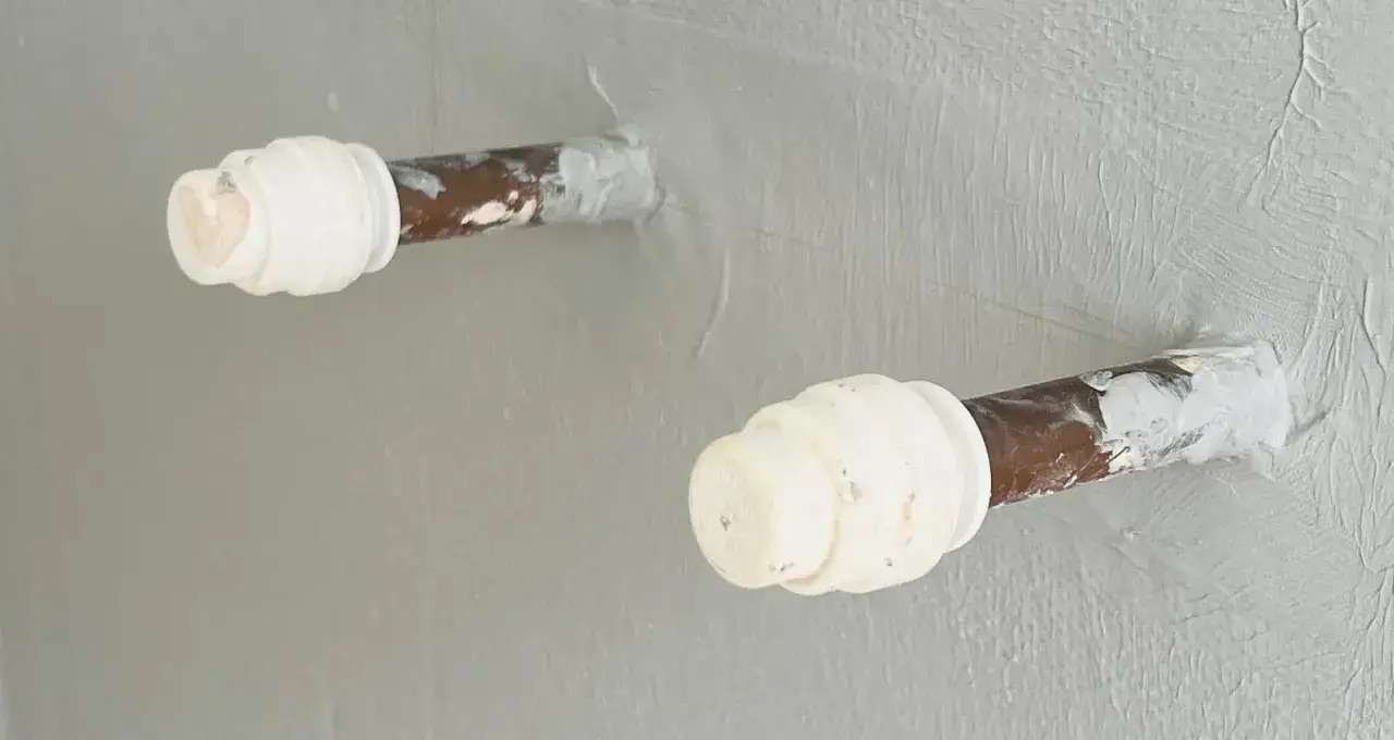 Dettaglio di tubi di rame che sporgono da una parete intonacata, con tappi bianchi a protezione. L'immagine mostra il risultato di un lavoro di forare piastrelle per tubi.