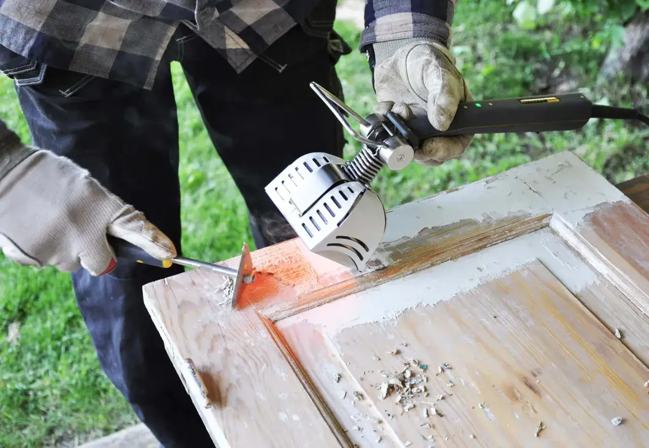 Come togliere la vernice dal legno: un professionista usa una pistola termica e una spatola per rimuovere strati di vernice da una porta in legno.