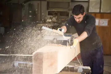Falegname usa pialla elettrica su un grosso tronco di legno, creando una nuvola di segatura. Il piallare significato è dare una finitura liscia.